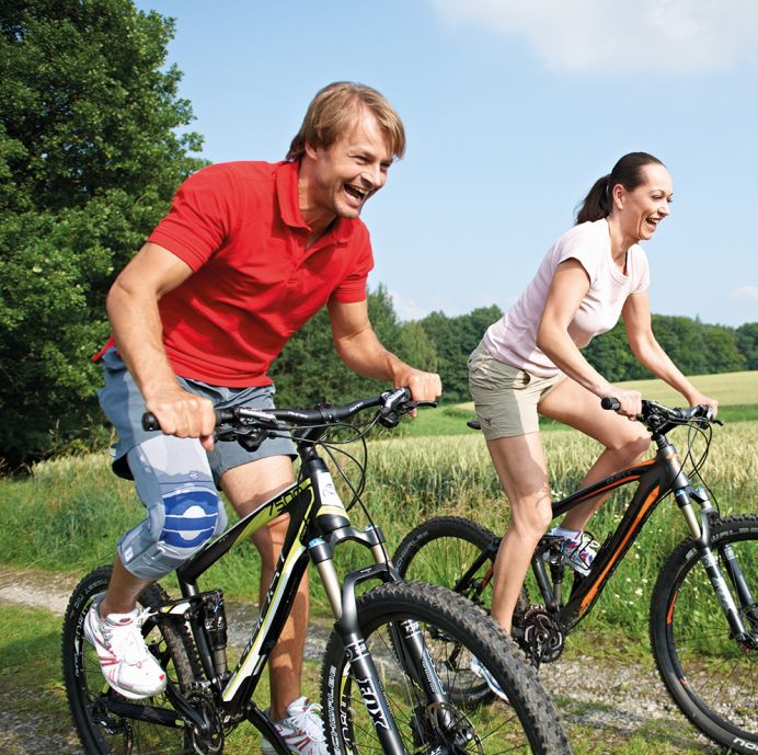 Mann und Frau fahren Fahrrad. Der Mann trägt eine Kniebandage. Beide lächeln. Hintergrund: Feld und blauer Himmel.