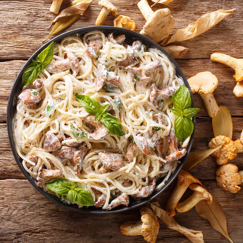 Teller Pasta mit Pilzen und Basilikum. Auf einem Holztisch, umgeben von Pilzen und Blättern.