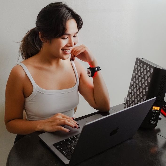 Frau mit Armband und Lichttherapiegerät. Sie sitzt an einem Tisch mit Laptop. Gerät strahlt rotes Licht.