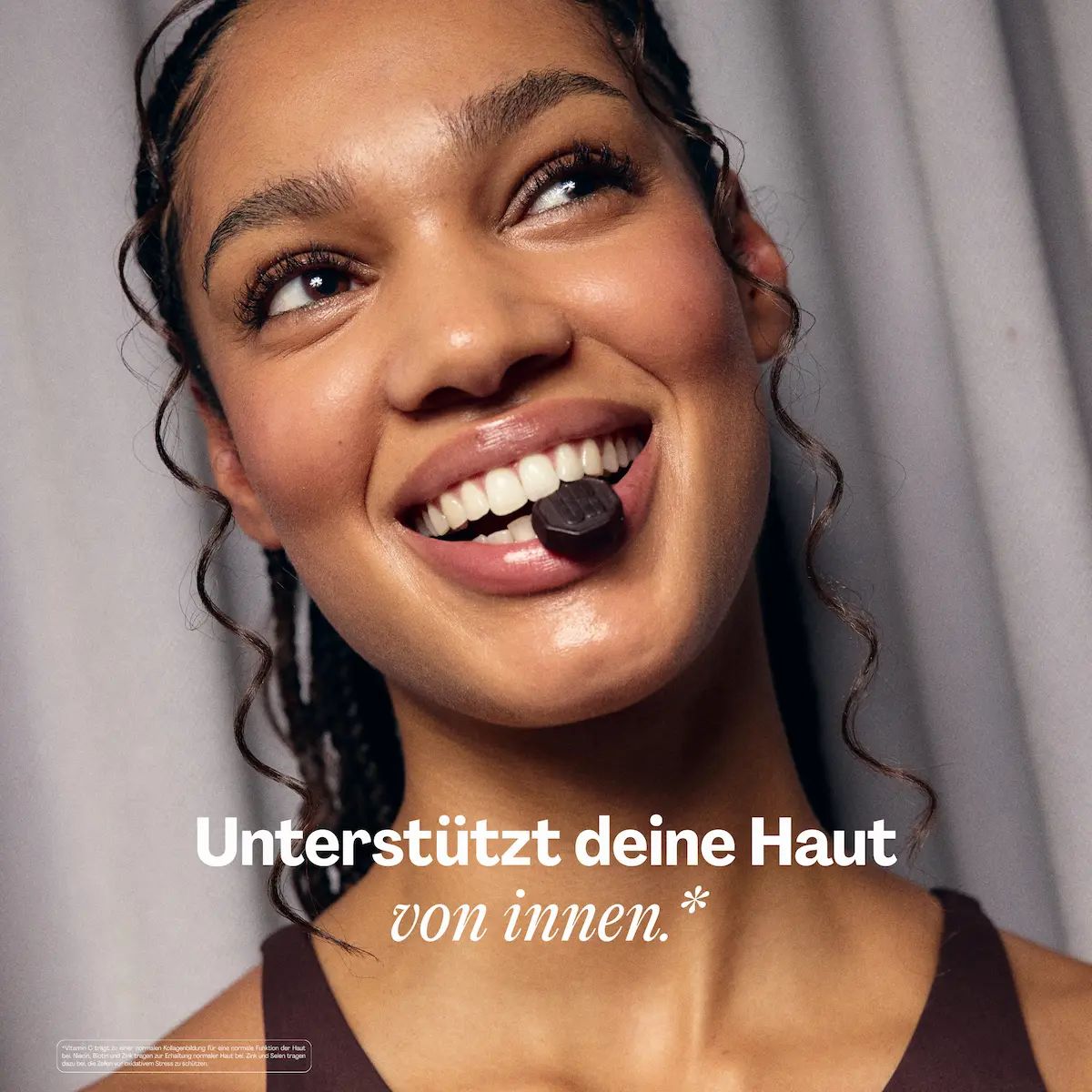 Eine Frau lächelt und hält eine Gummibärchen im Mund. Text: Unterstützt deine Haut von innen.