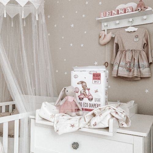 Windelpackung auf Wickeltisch. Daneben Stofftier-Hase und Babykleidung. Hintergrund: Sternenmuster an Wand.