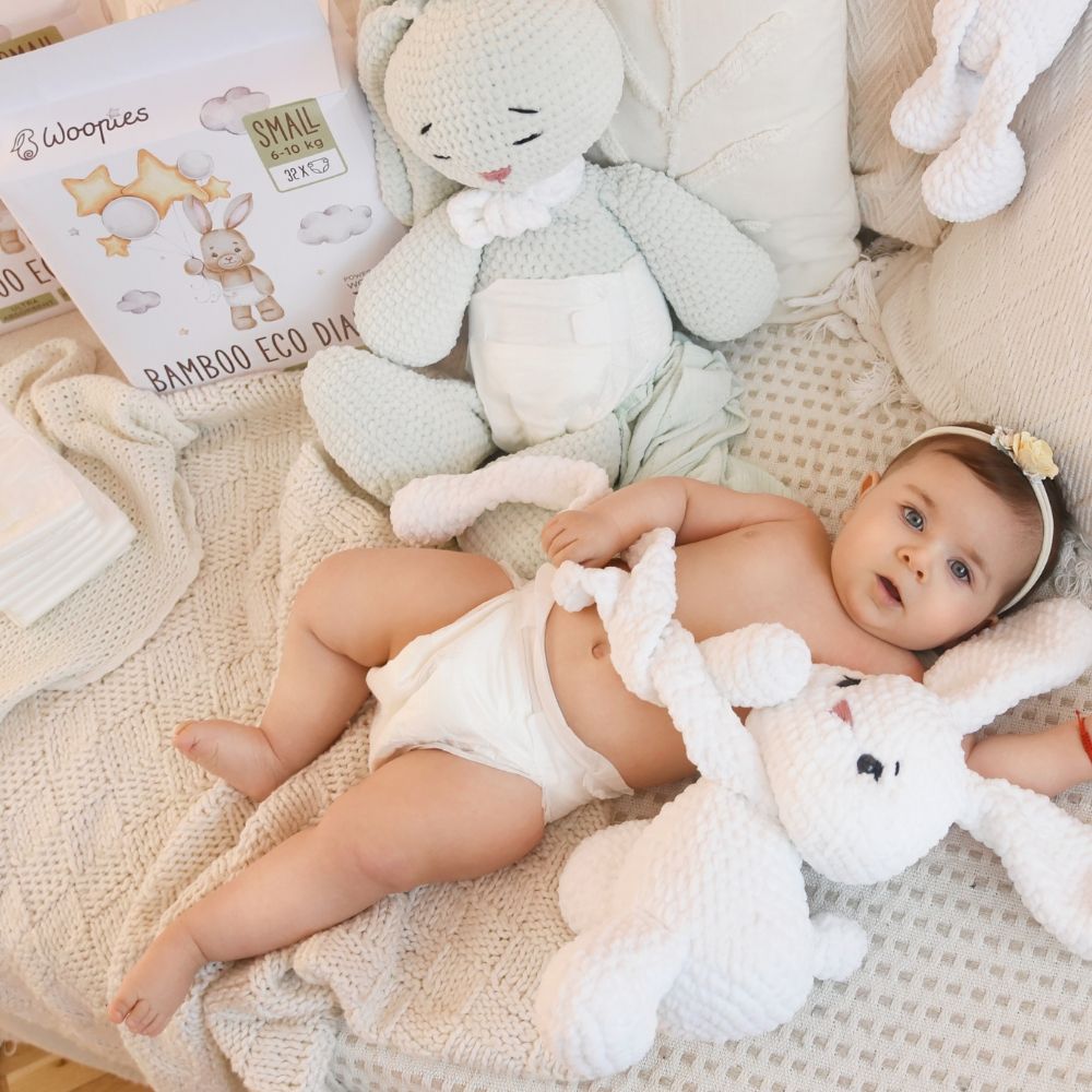 Baby liegt auf dem Bett, umgeben von Spielzeug. Im Hintergrund: Packung Babywindeln. Aufdruck: Hase mit Luftballons, Sterne, Wolken. Text: Bamboo Eco Diapers, SMALL.