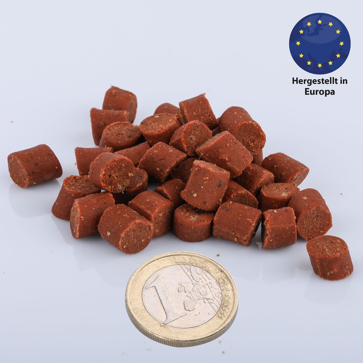 Haufen brauner, zylindrischer Snacks. Daneben eine 1-Euro-Münze. Oben rechts: EU-Siegel „Hergestellt in Europa“.