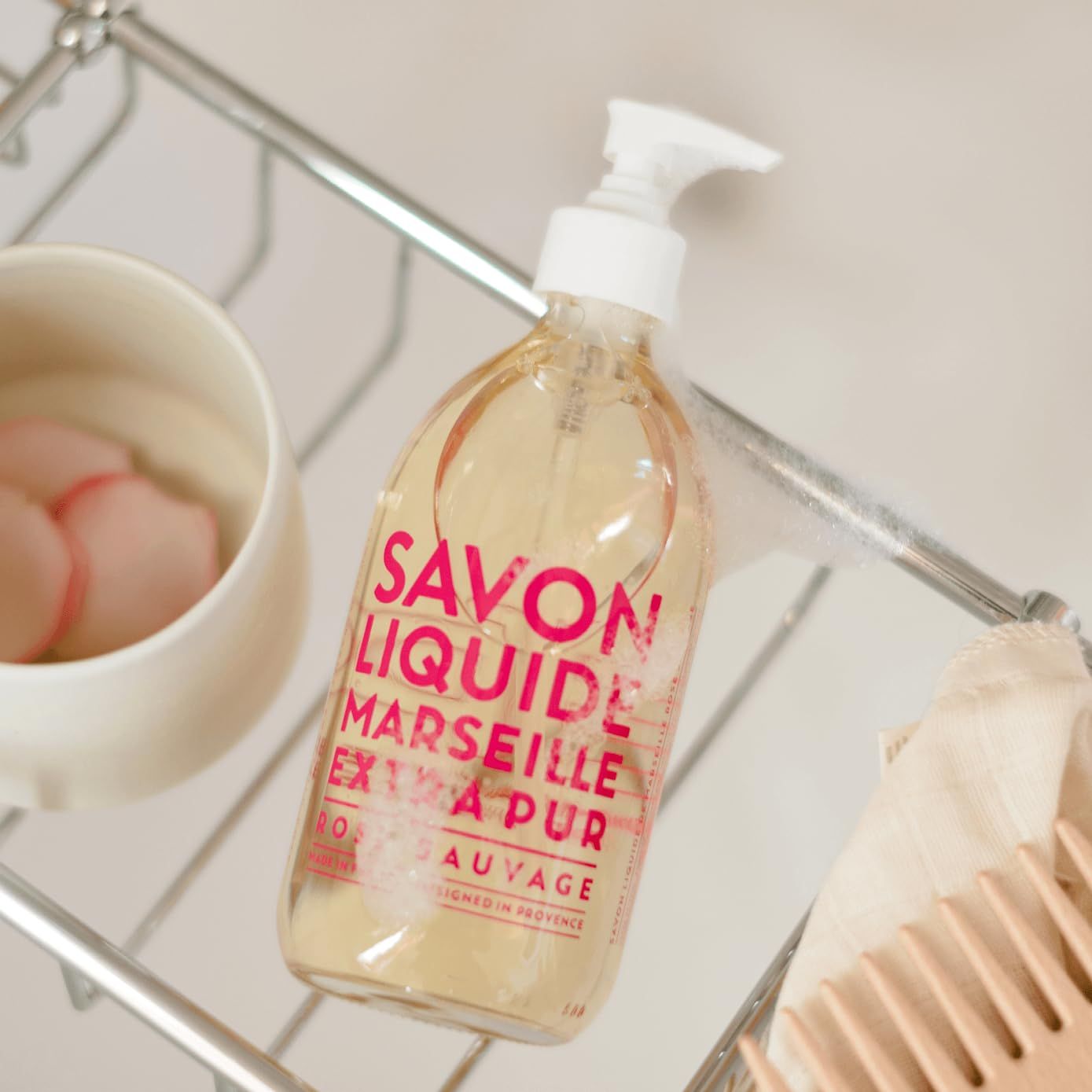 Flüssigseife mit Pumpe. Aufschrift: Savon Liquide Marseille Extra Pur Rose Sauvage. Flasche auf einem Regal, neben einer Schüssel und einem Kamm.