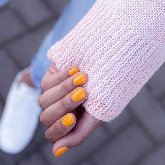 Hand mit orange lackierten Nägeln. Hellrosa Pullover und weiße Schuhe im Hintergrund.