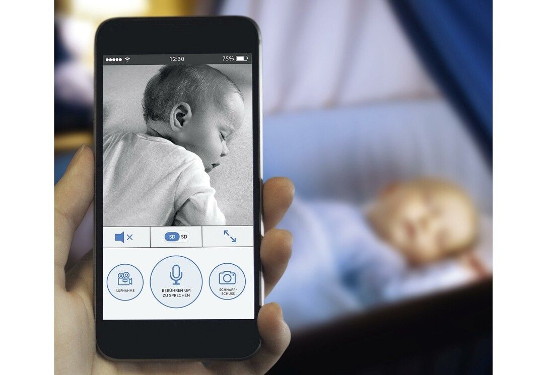 Smartphone mit Baby-Video. Auf dem Bildschirm sind Bedienelemente zu sehen. Im Hintergrund ein Baby in einer Wiege.