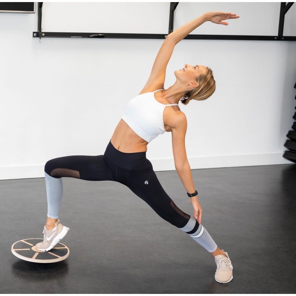 Frau auf Balanceboard in Yoga-Pose. Board mit schwarzer, rutschfester Oberfläche. Frau in Sportkleidung, Arm erhoben.
