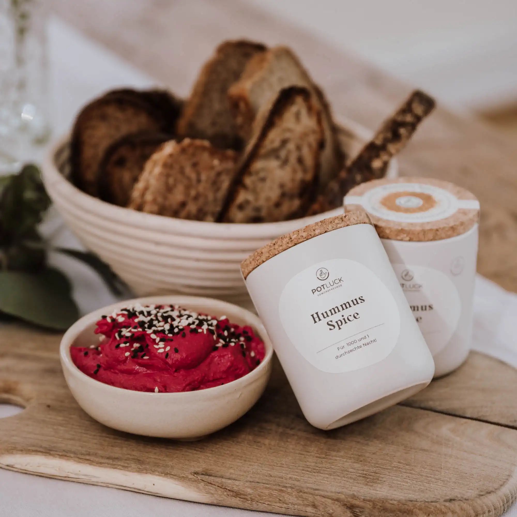 Zwei weiße Behälter mit Korkdeckel. Aufkleber "Hummus Spice". Dazu Brot und Hummus in Schalen auf Holzbrett.