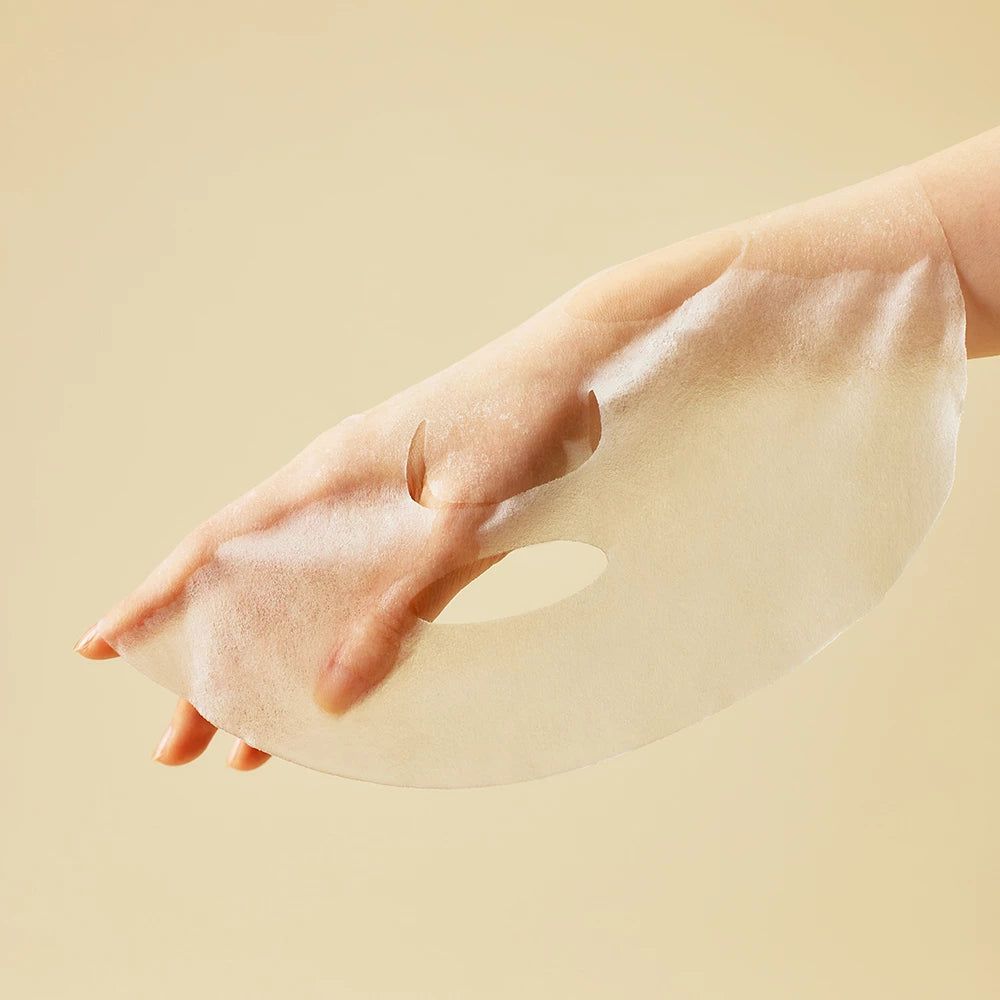 Gesichtsmaske in einer Hand gehalten. Die Maske hat Löcher für Augen und Mund. Heller Hintergrund.