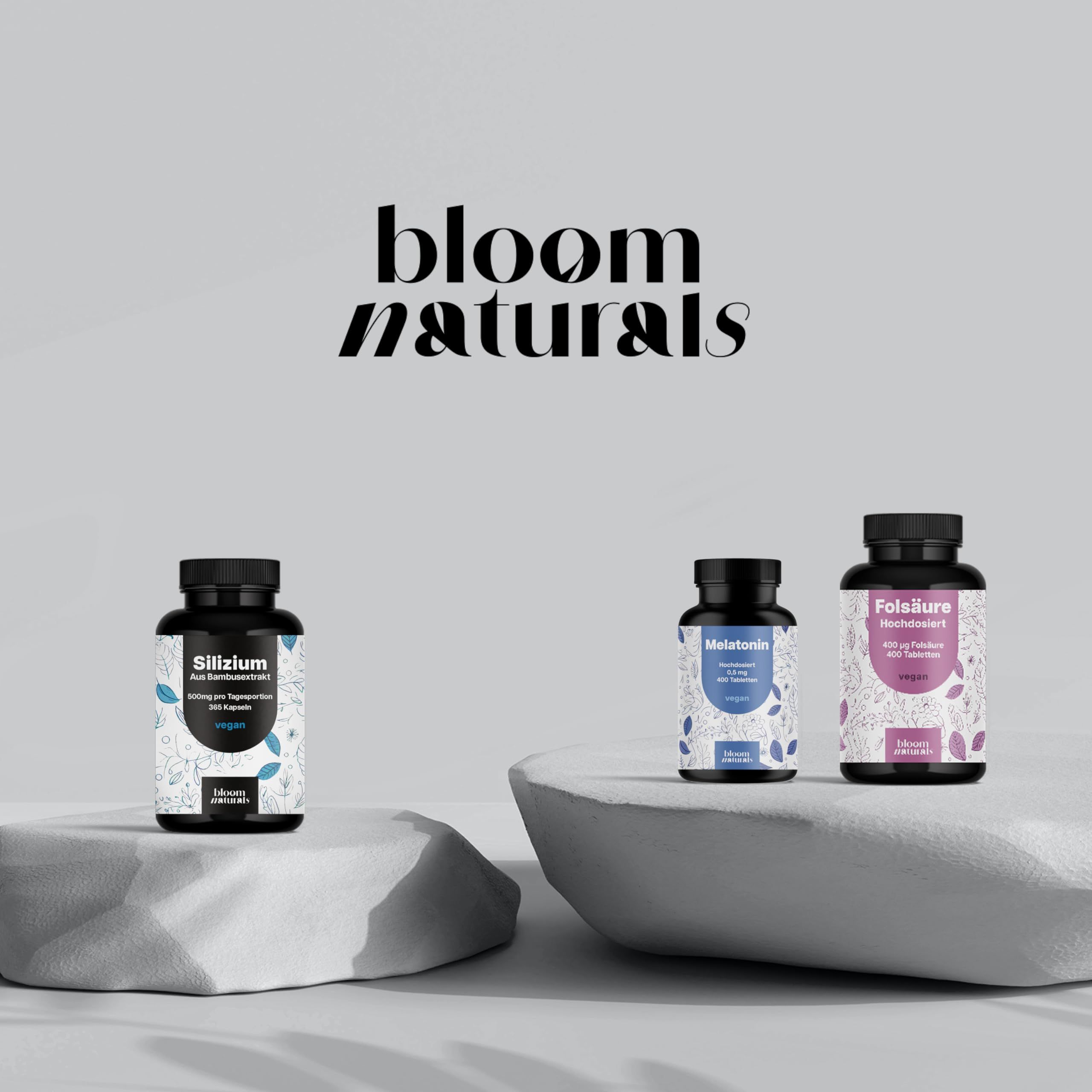 Drei Dosen Bloom Naturals Produkte: Silizium, Melatonin, Folsäure auf Steinen.
