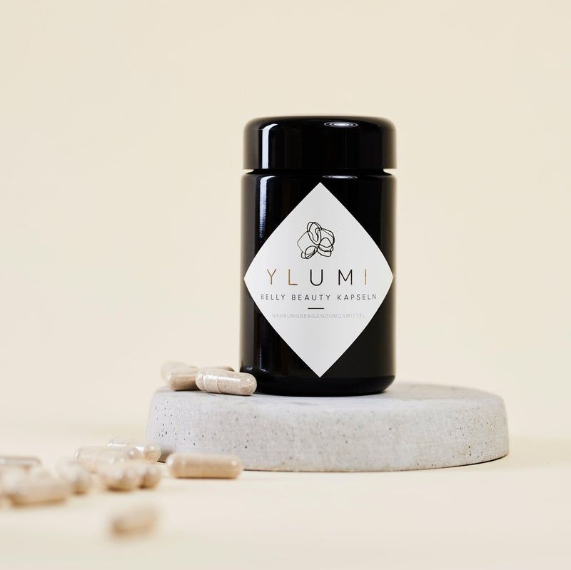 Schwarze Dose mit Etikett. YLUMI BELLY BEAUTY KAPSELN. Auf Podest. Kapseln daneben. Heller Hintergrund.