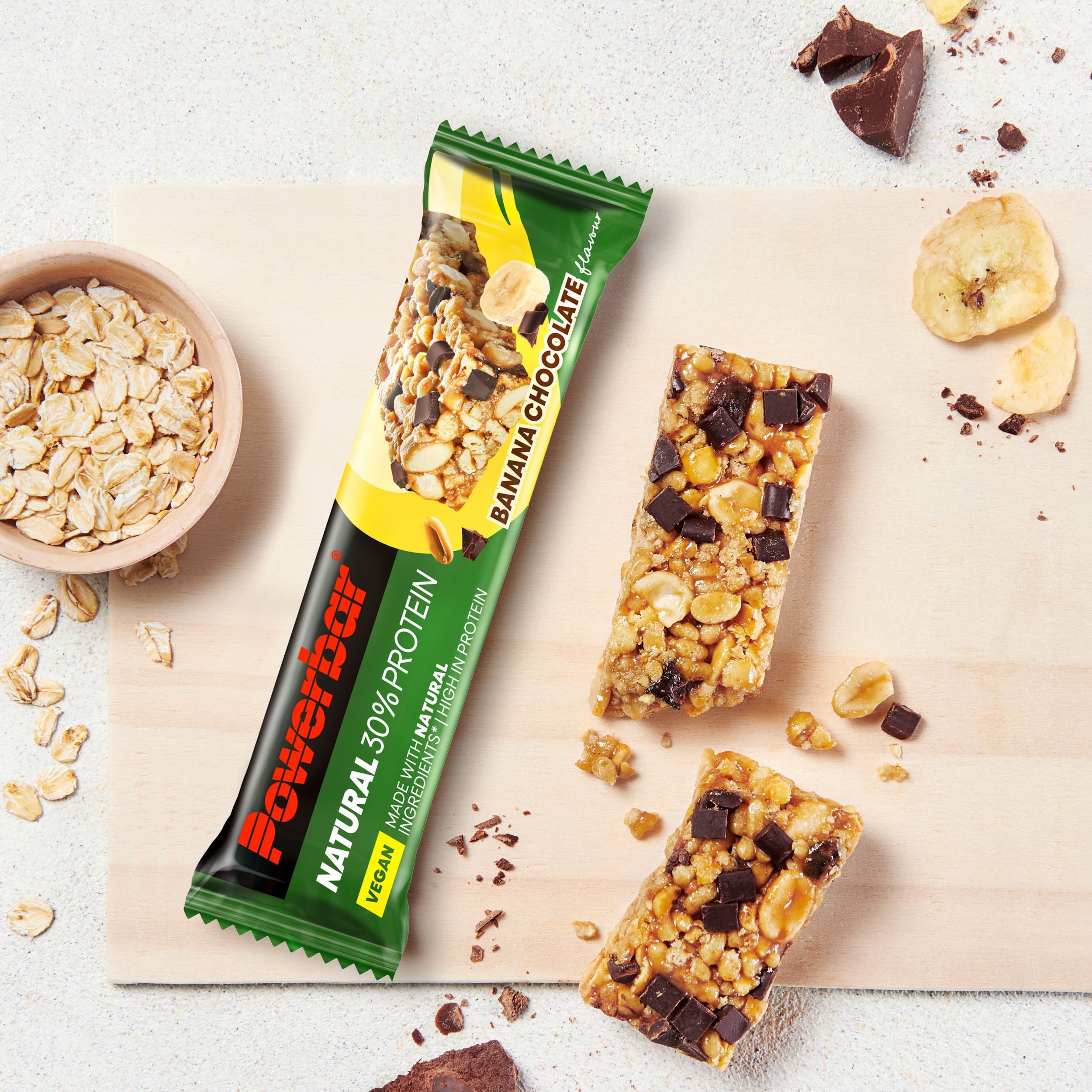 POWERBAR Riegel Banana Chocolate, teilweise zerbrochen, auf Holzbrett. Daneben Haferflocken und Bananenscheiben. Vegan, mit 30% Protein.