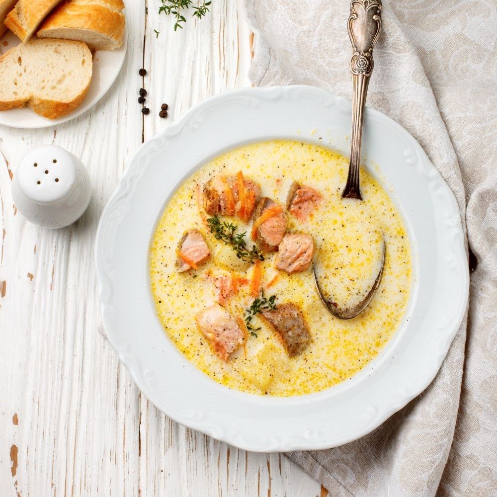 Teller mit cremiger Suppe, Lachs, Gemüse und Kräutern. Dazu Löffel, Salzstreuer und Brot auf Holzgrund.
