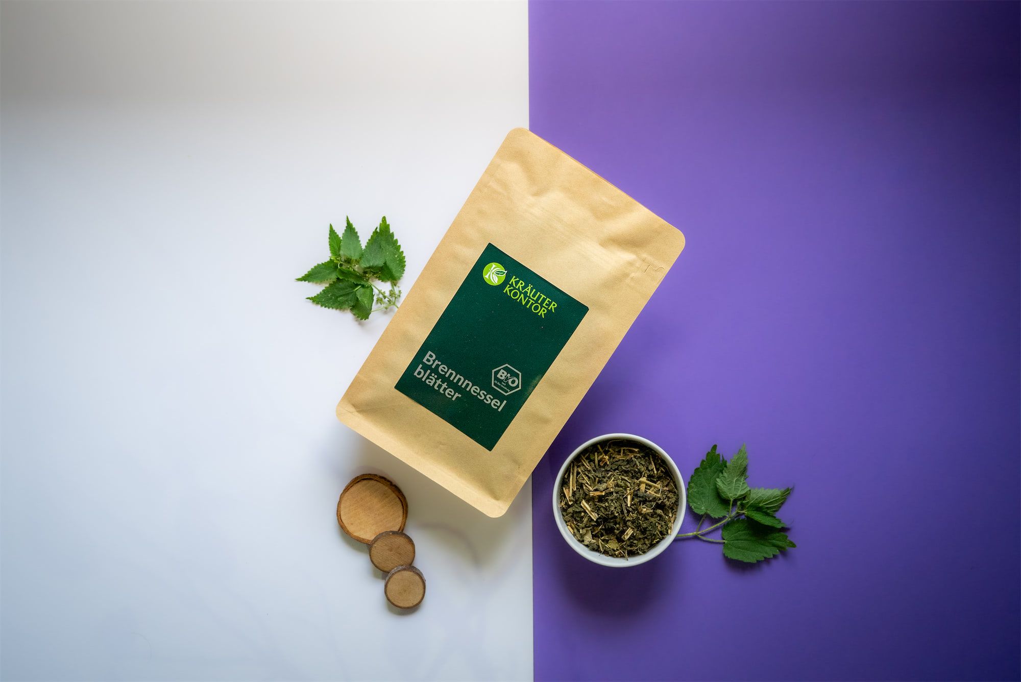 Kräuterkontor Brennnesselblätter-Packung, umgeben von Kräutern, Holzscheiben und einer Schale mit Kräutern. Hintergrund zweifarbig.