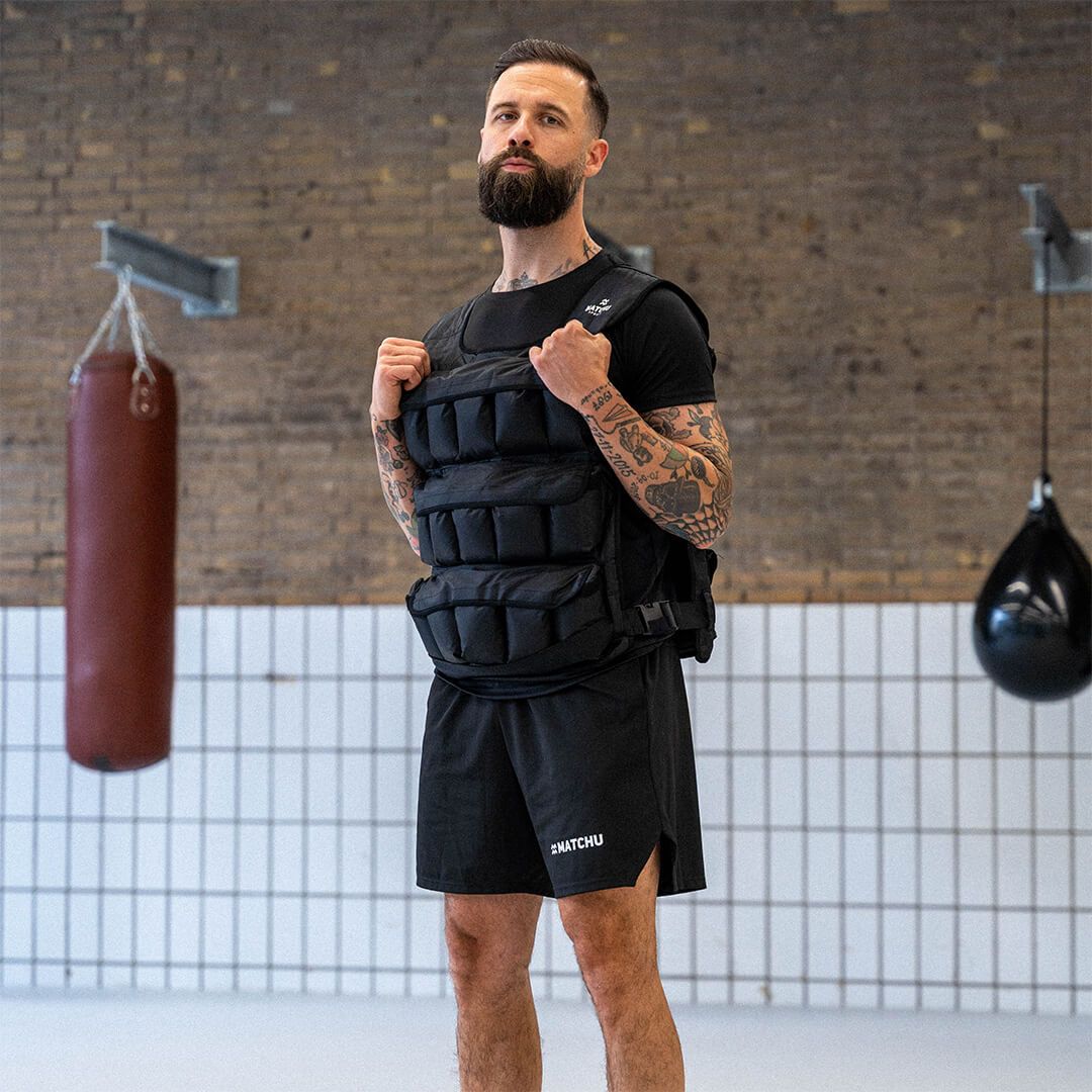 Mann mit schwarzer Gewichtsweste. Marke Matchu Sports. Hintergrund: Fitnessstudio mit Boxsäcken.