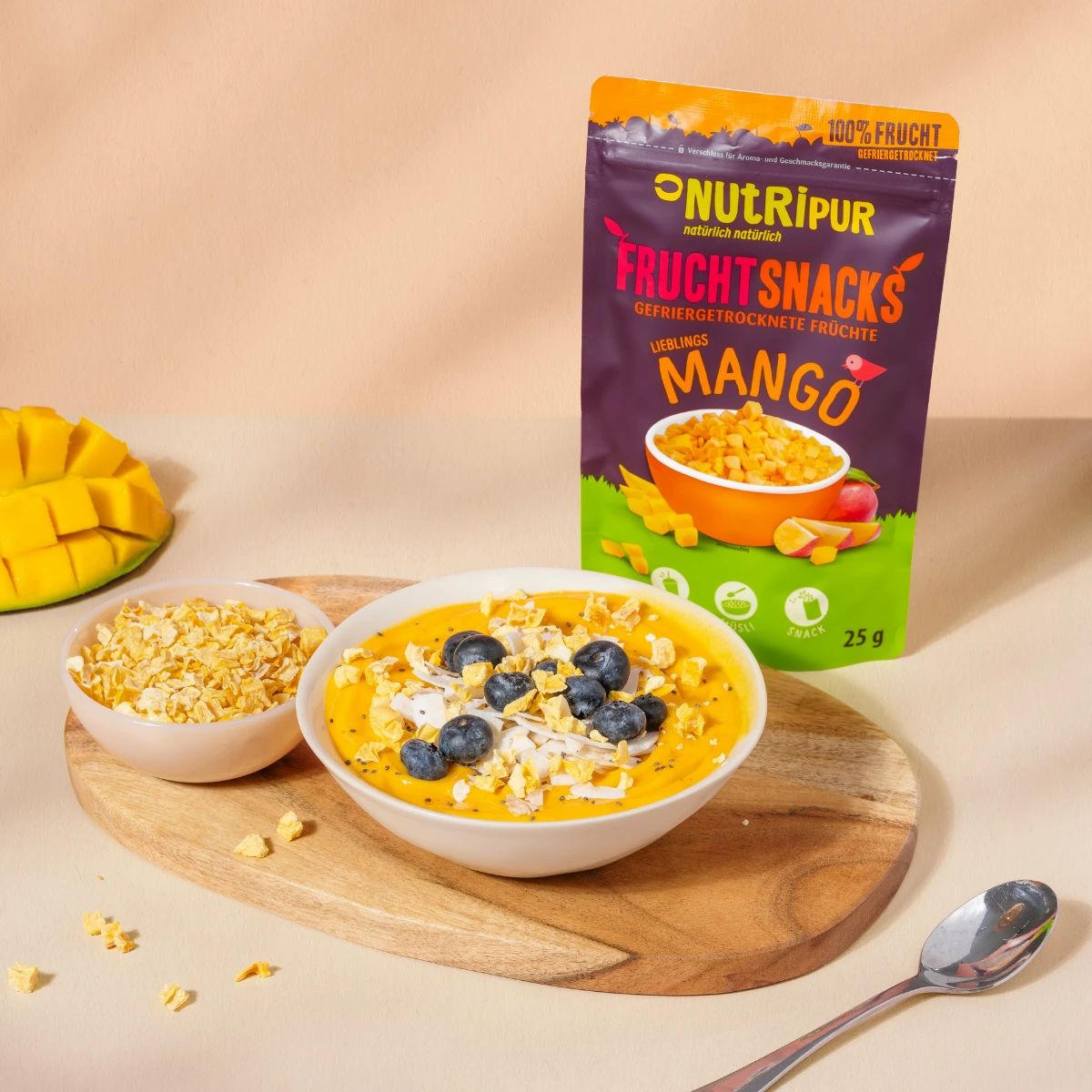 Mango-Fruchtsnacks in Schalen mit Blaubeeren. Daneben die Verpackung von NutriPur Mango-Fruchtsnacks. Auf einem Holzbrett und einem Löffel.