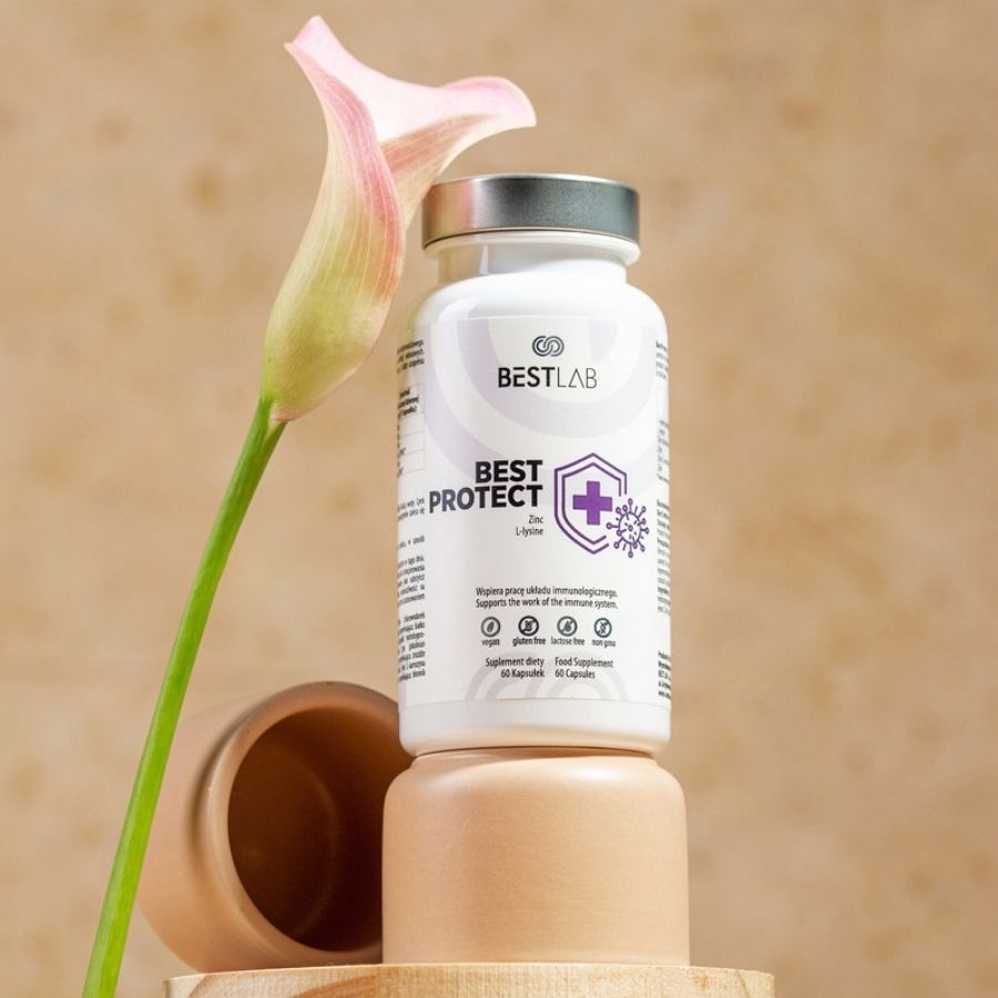 Weiße Flasche mit silbernem Deckel, platziert auf einem Holzsockel. Aufschrift: BEST LAB, BEST PROTECT, Zink L-Lysin. Dekoration: Blume und Keramikgefäß.