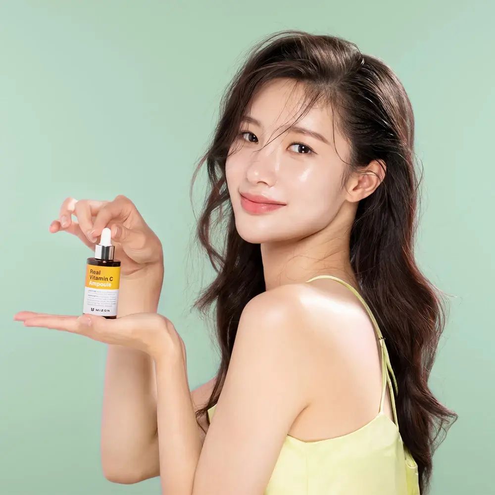 Frau hält eine kleine Flasche Real Vitamin C Ampoule. Gelbes Top, grüner Hintergrund. Marke: MIZON.