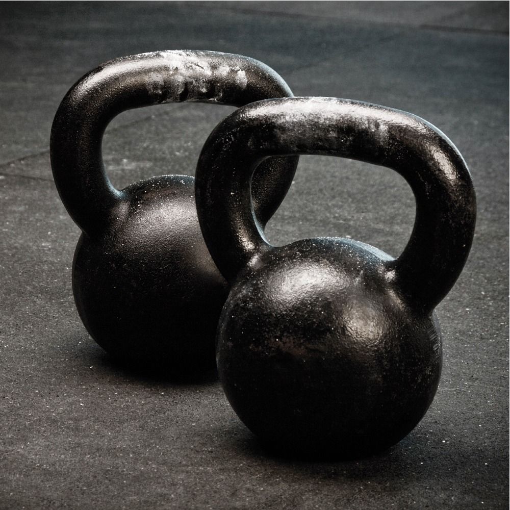 Zwei schwarze Kettlebells aus Gusseisen mit Griffen, auf dunklem Boden.
