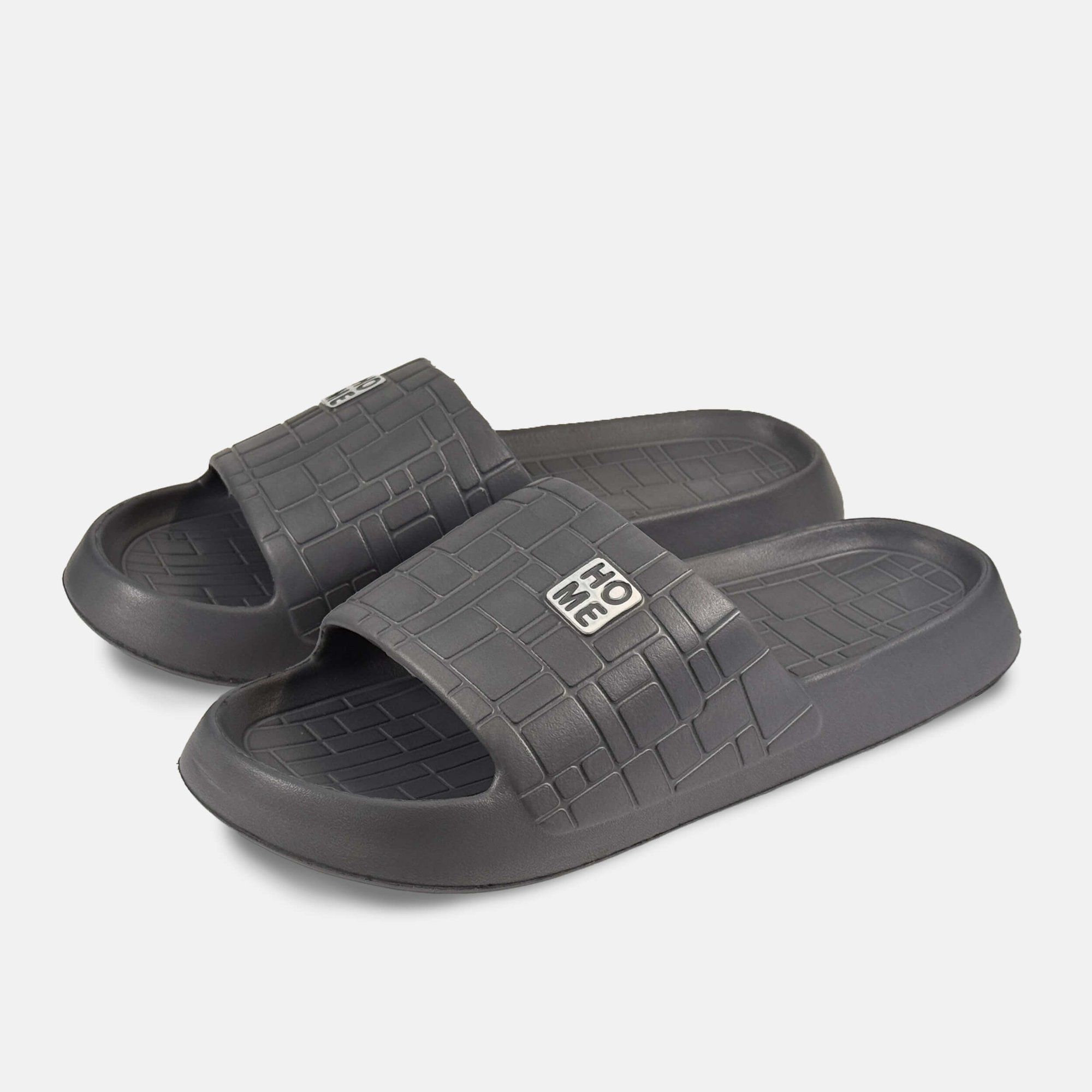 Graue Pantoletten mit quadratischem Muster und "HOME"-Logo. Breite Form. Zwei Schuhe nebeneinander.