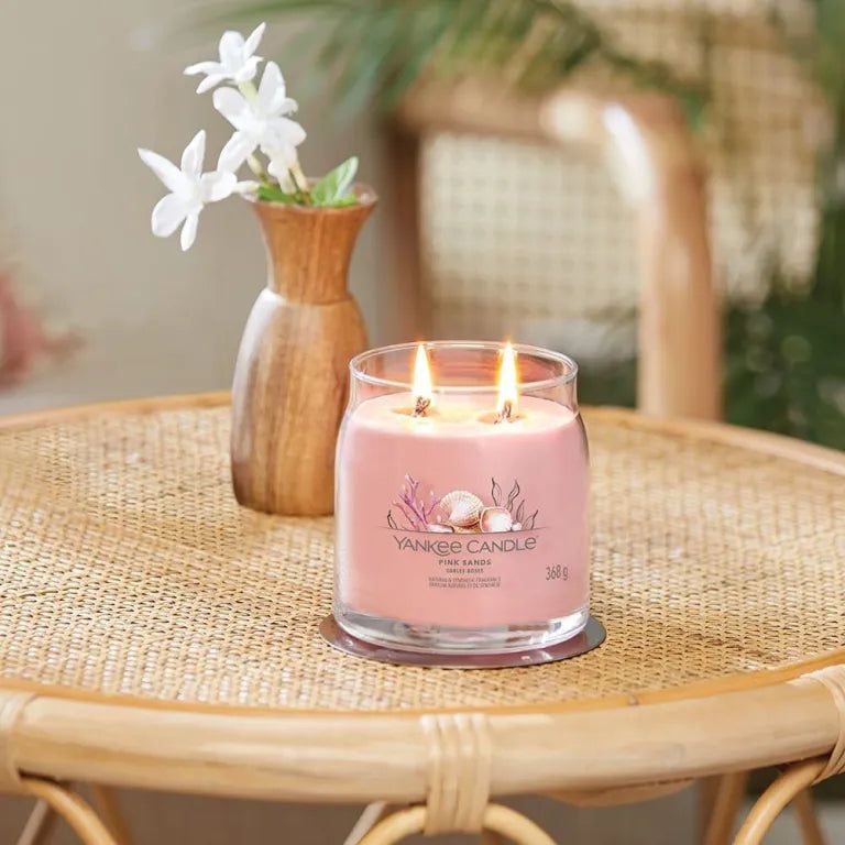 Rosa Kerze Yankee Candle Pink Sands auf einem Tisch. Zwei Dochte brennen. Vase mit Blumen im Hintergrund.
