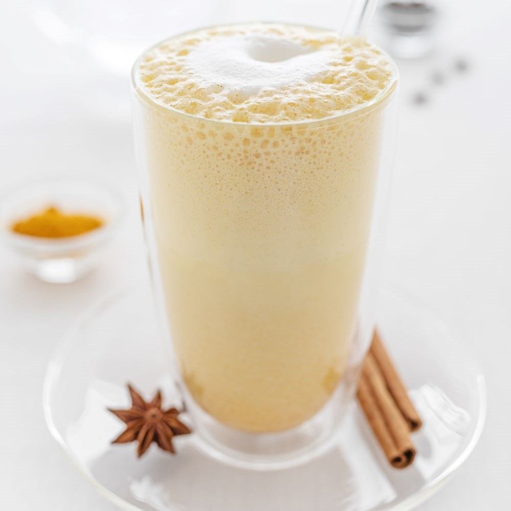 Hohes Glas mit goldener Milch und Schaum. Zimtstange, Anisstern. Kleines Schälchen mit Kurkuma.