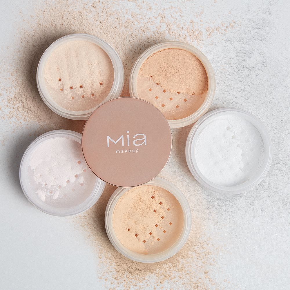 Diversi contenitori di cipria Mia Makeup. Sei contenitori sono parzialmente aperti, rivelando ciprie di diverse tonalità. Il coperchio di un contenitore è appoggiato accanto. Sul coperchio si legge "mia makeup".