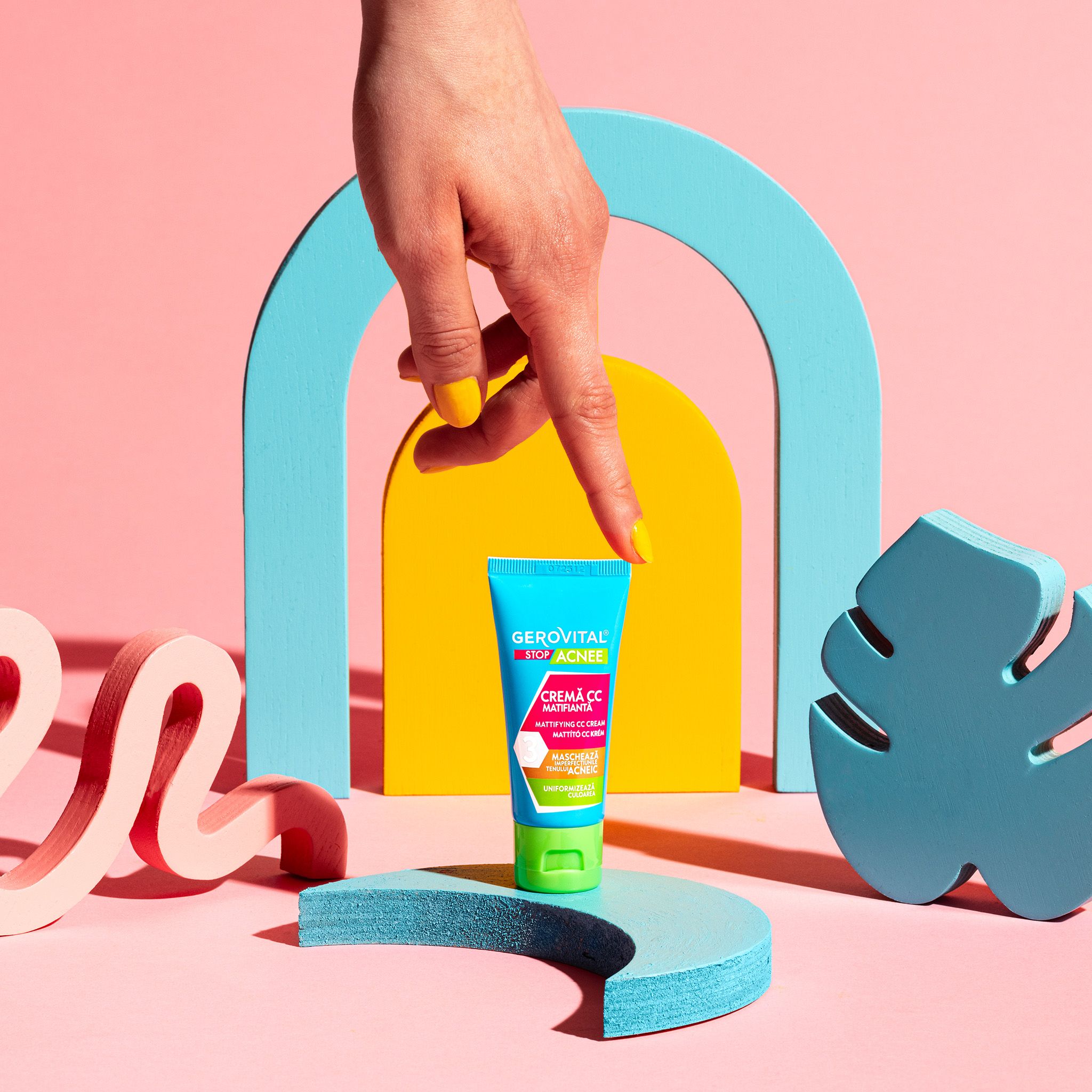 Gerovital Stop Acnee CC Creme Tube auf einem Podest. Türkis-grüne Tube mit grünem Deckel. Gelbe Hand.