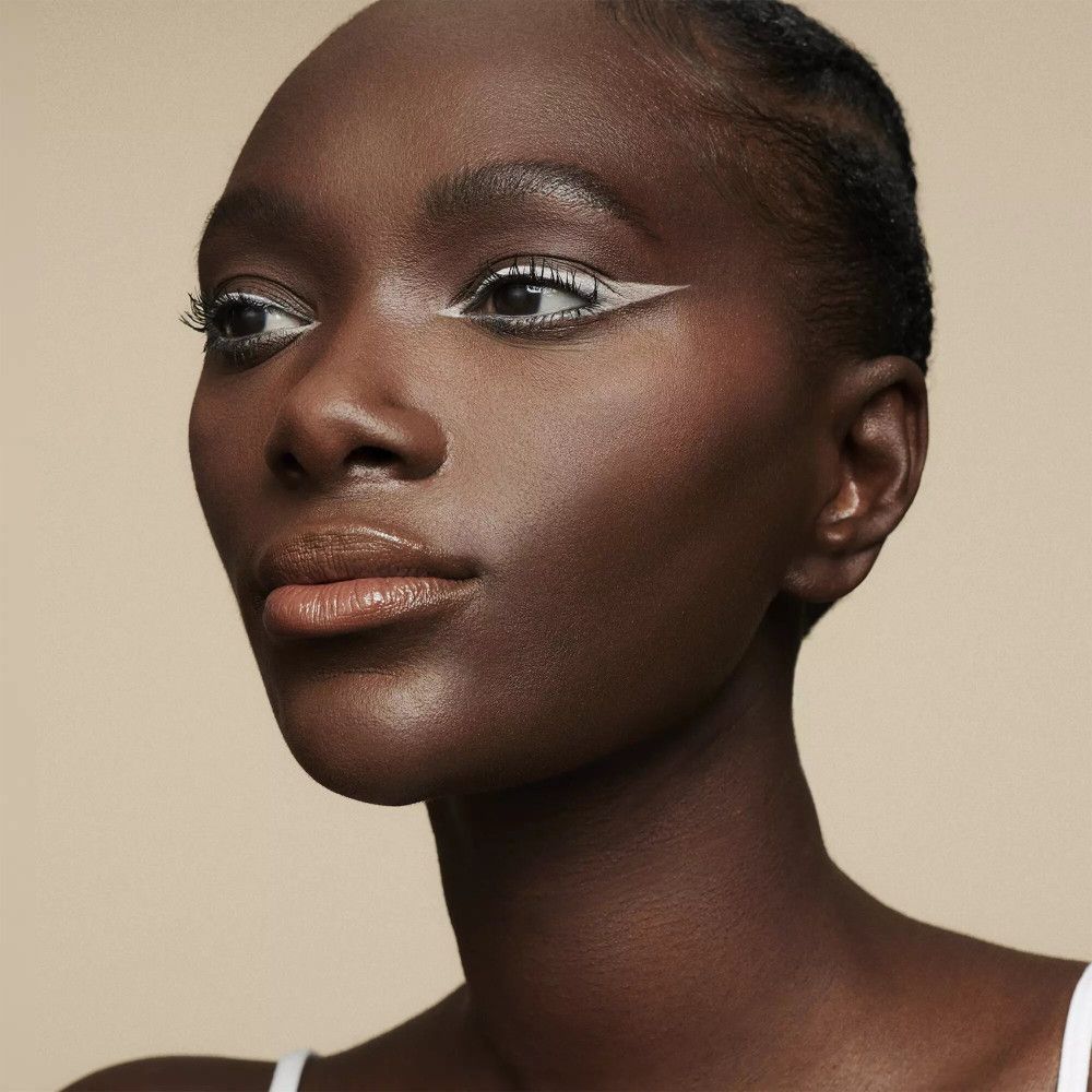 Nahaufnahme einer Frau mit dunkler Haut, die weißen Eyeliner trägt. Ihre Lippen sind leicht geschminkt. Der Hintergrund ist einfarbig.