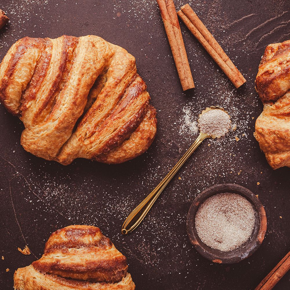 Franzbrötchen, Zimtstangen, Gewürz in Schale und Löffel. Draufsicht auf dunklem Untergrund. Zucker gestreut.