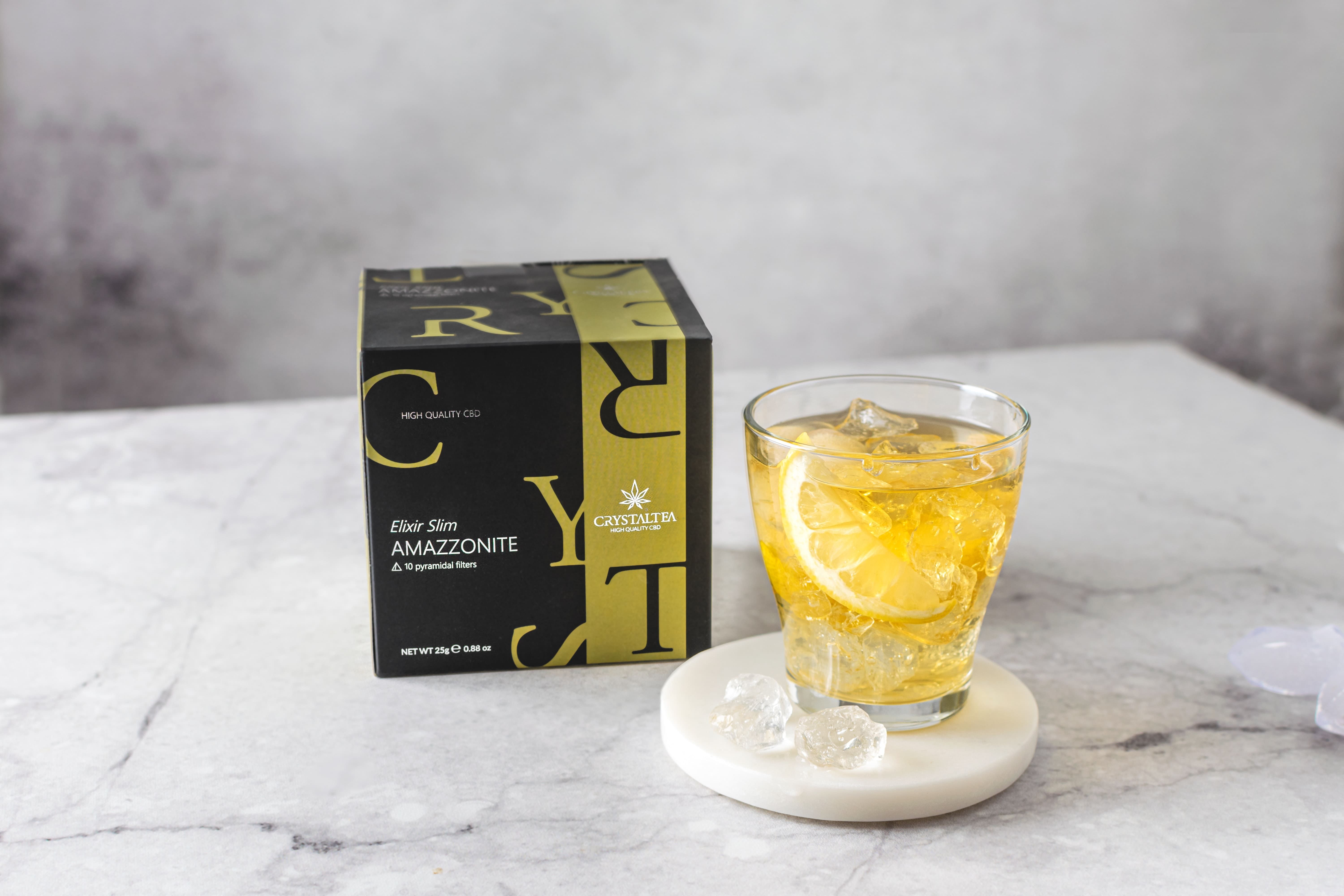 Glas mit Eiswürfeln und Zitronenscheibe. Daneben eine Schachtel mit Produktnamen und Logo.
