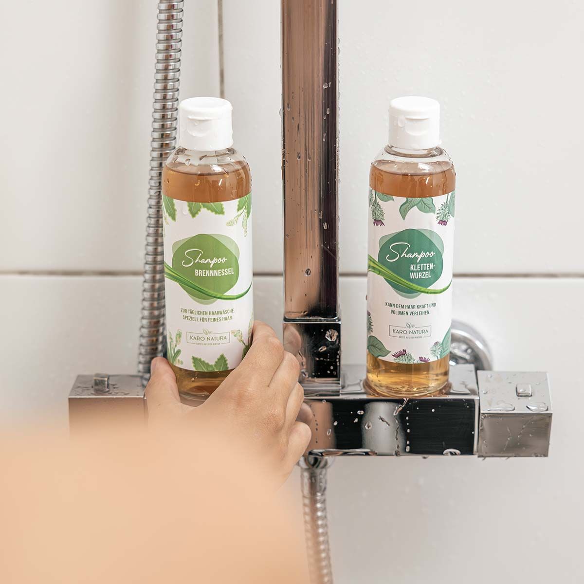 Zwei Flaschen Shampoo auf einer Ablage in einer Dusche. Etiketten mit Schriftzügen. Karo Natura Logo.