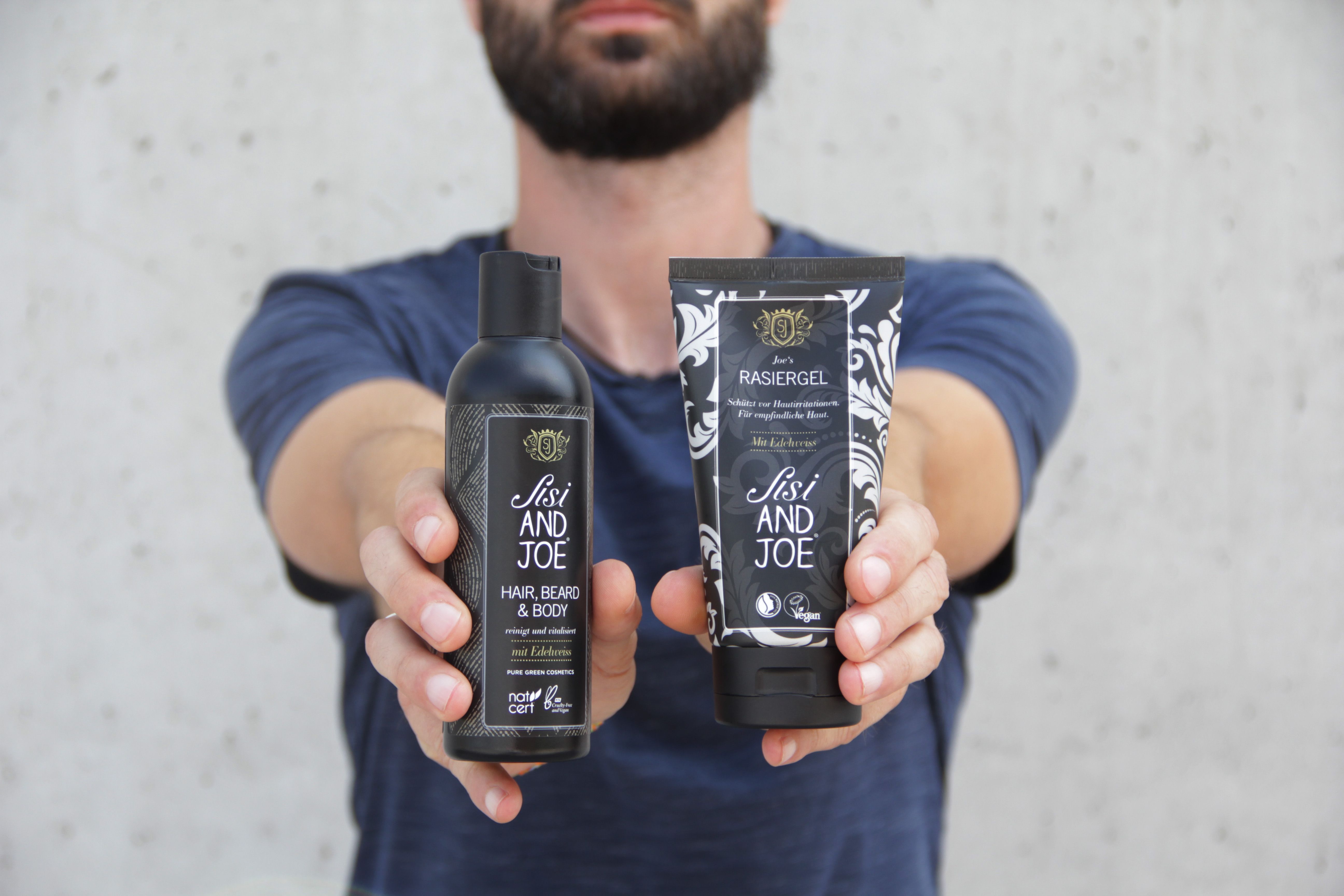 Mann hält zwei schwarze Flaschen. Links: Hair, Beard & Body Wash. Rechts: Rasiergel. Beide mit Logo und Text.