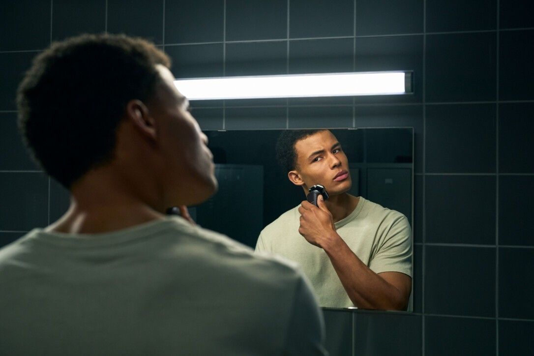 Mann rasiert sich vor einem Spiegel mit einem elektrischen Rasierer. Helles T-Shirt.