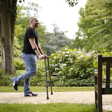 Mann geht im Park mit zwei Unterarmgehhilfen. Er trägt Jeans, ein schwarzes T-Shirt und Turnschuhe.