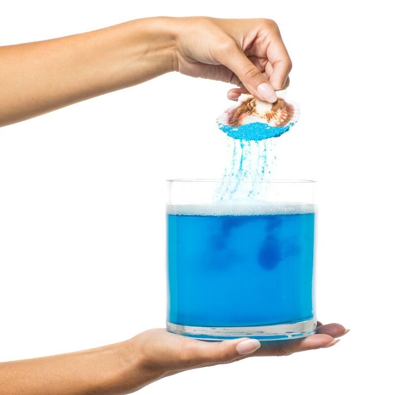 Hände halten Glas mit blauem Wasser. Muschel wird über Glas gehalten, Salz rieselt hinein. Wasser mit Schaum.