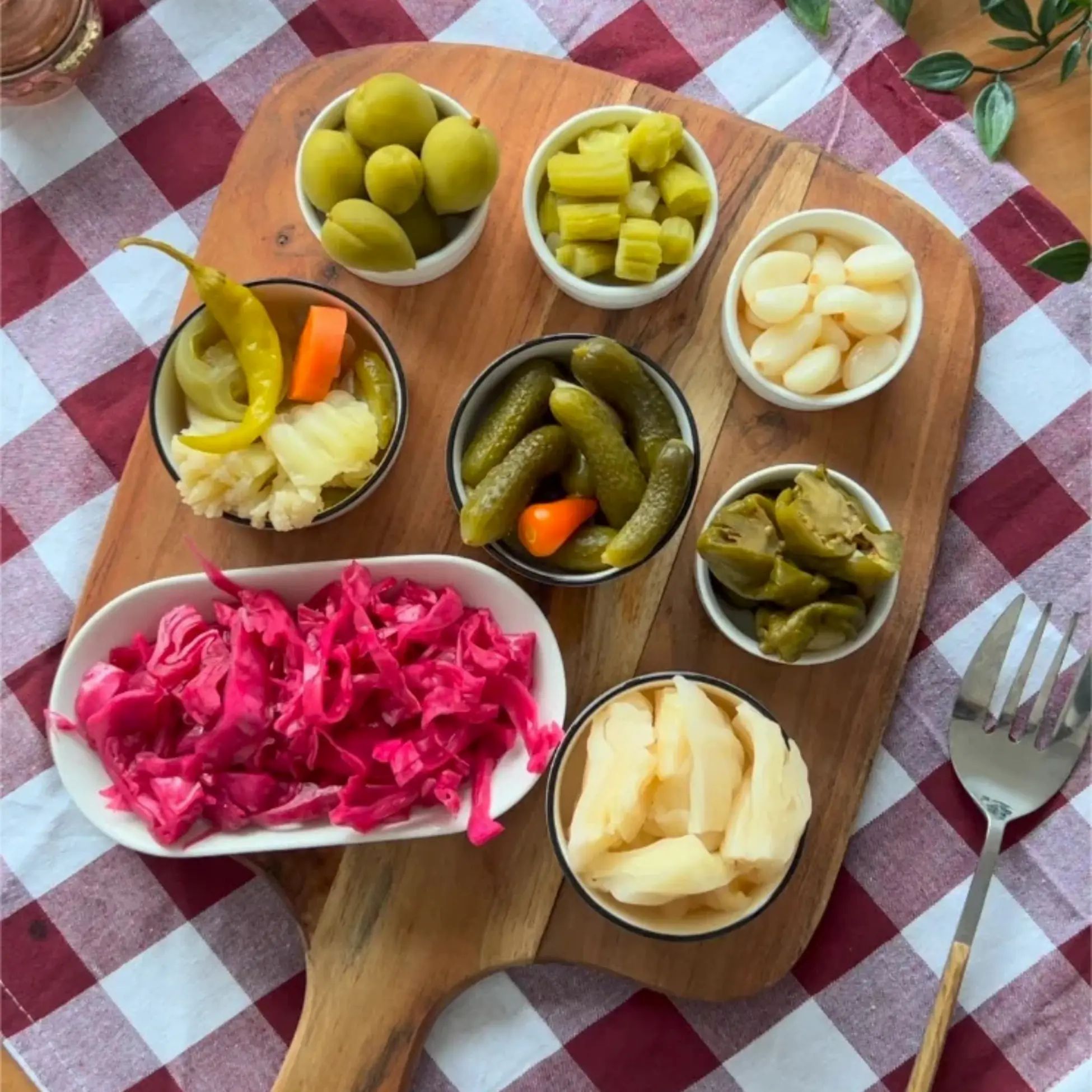Holzbrett mit verschiedenen eingelegten Gemüsesorten in kleinen Schalen. Darunter Oliven, Gurken, Paprika und Kraut.