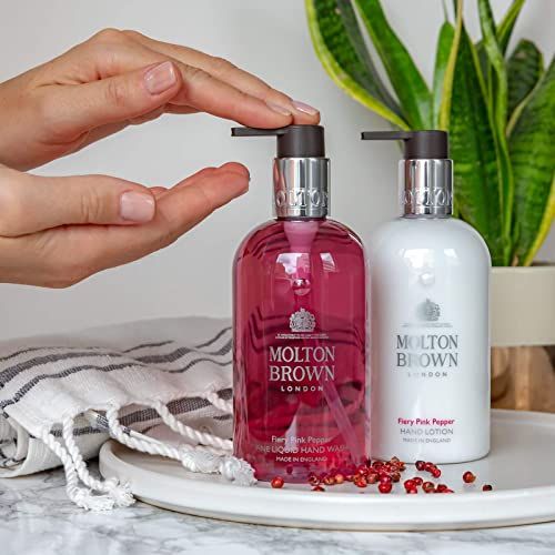Hände drücken auf eine Pumpe. Zwei Molton Brown Handseifen. Auf einem Tablett. Handtuch. Rosa Pfefferkörner. Pflanze im Hintergrund.