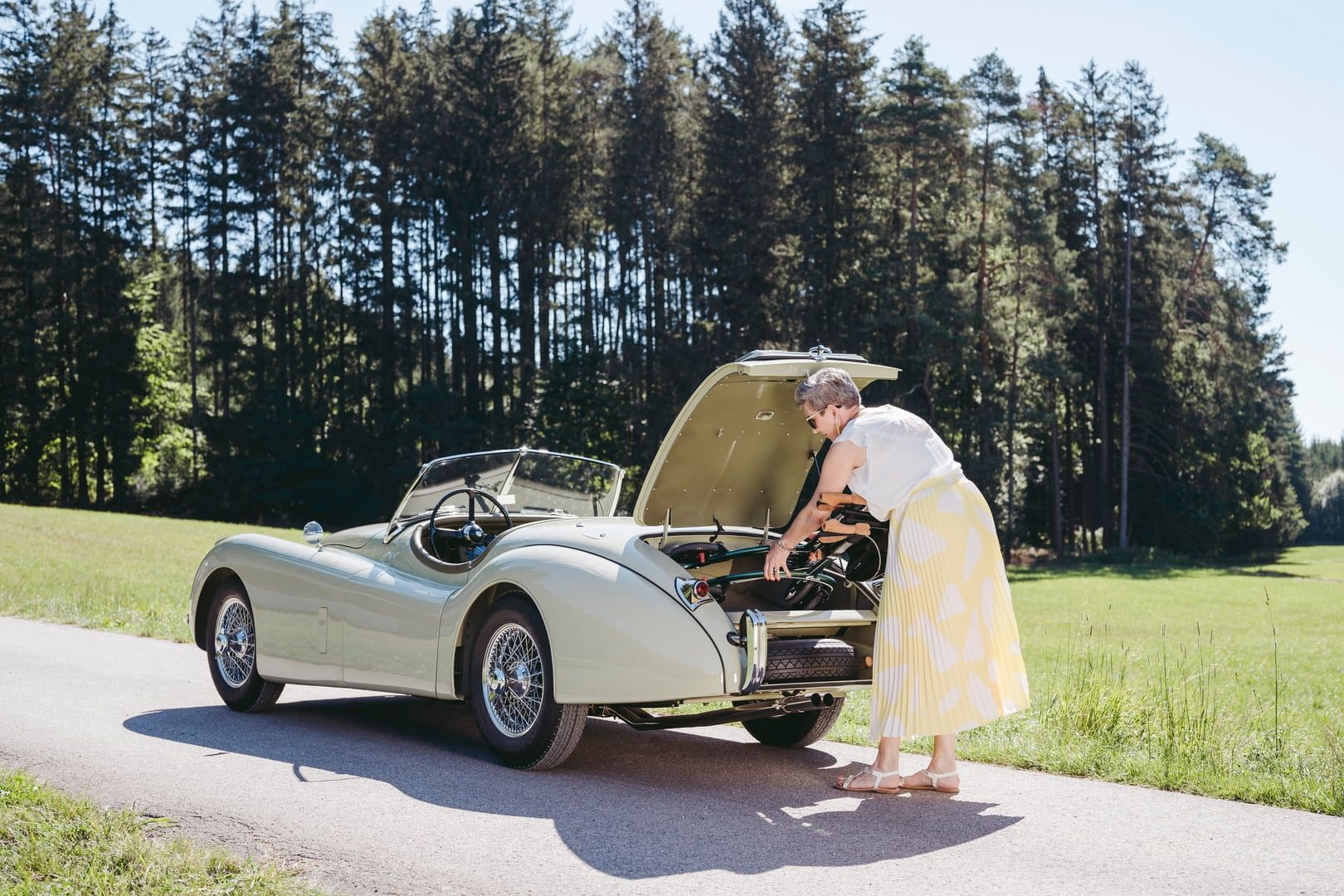 Ältere Frau öffnet die Motorhaube eines Oldtimers. Sie trägt einen gelben Rock und ein weißes Oberteil. Hintergrund: Straße.