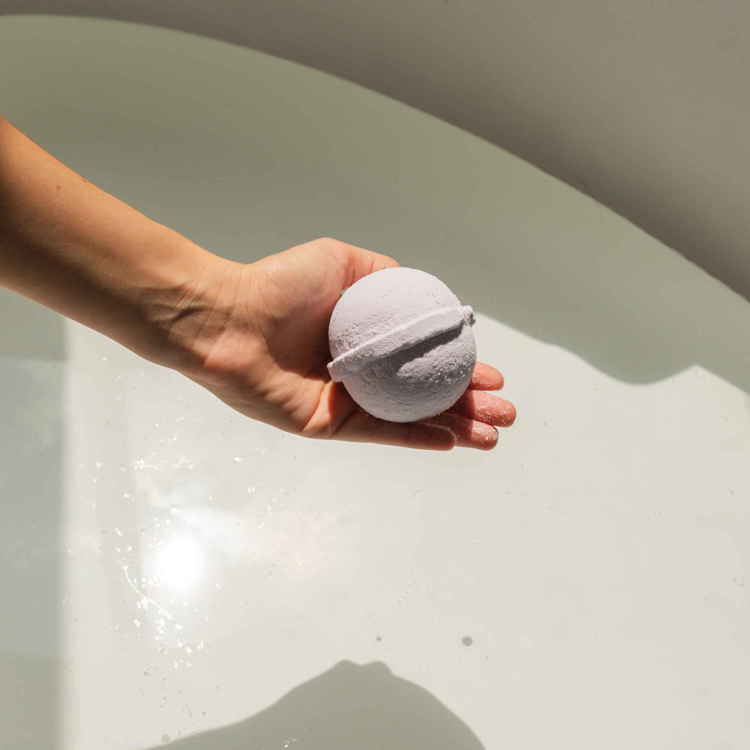 Hand hält eine rosa Badekugel über Badewasser. Gerillte Oberfläche sichtbar.
