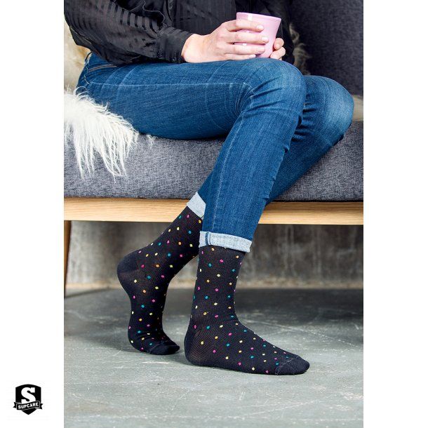 Person trägt schwarze Socken mit bunten Punkten. Marke SupCare am Knöchel. Sitzt auf Couch.