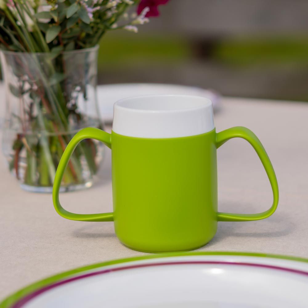 Grüner Becher mit zwei Griffen und weißem Rand. Steht auf einem Tisch. Im Hintergrund eine Vase mit Blumen und ein Teller.
