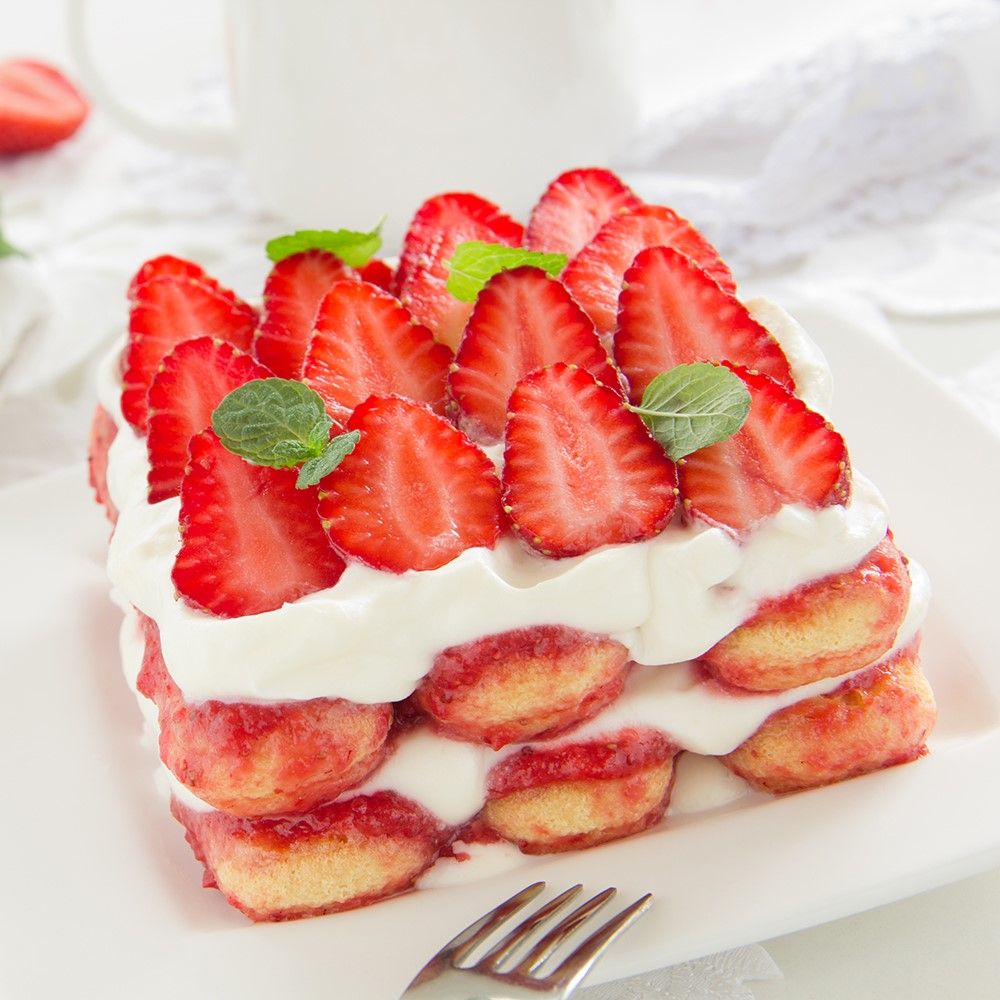 Rechteckiger Kuchen mit Erdbeeren und Sahne. Dekoriert mit Minze. Auf weißem Teller mit Gabel.