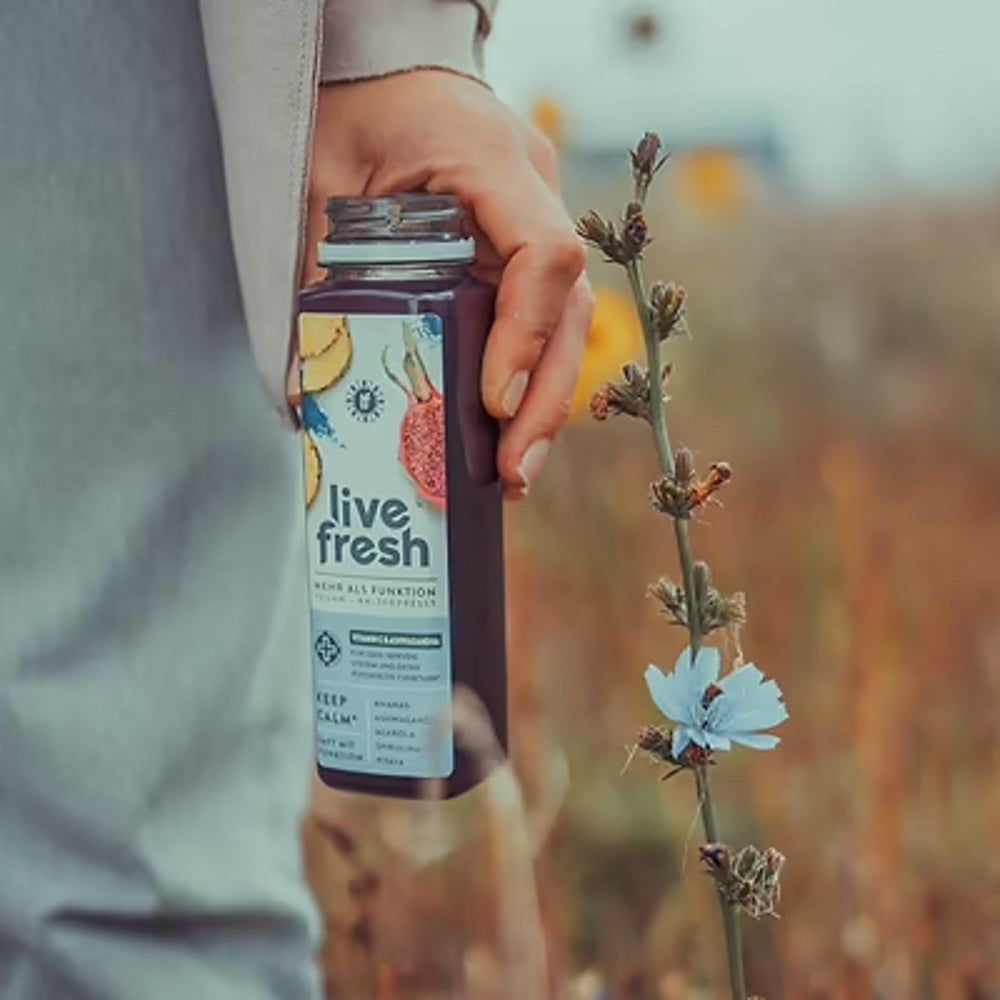 Eine Hand hält eine Flasche LiveFresh Saft. Hintergrund: Blumen und Feld. Etikett mit Zutaten und Logo.
