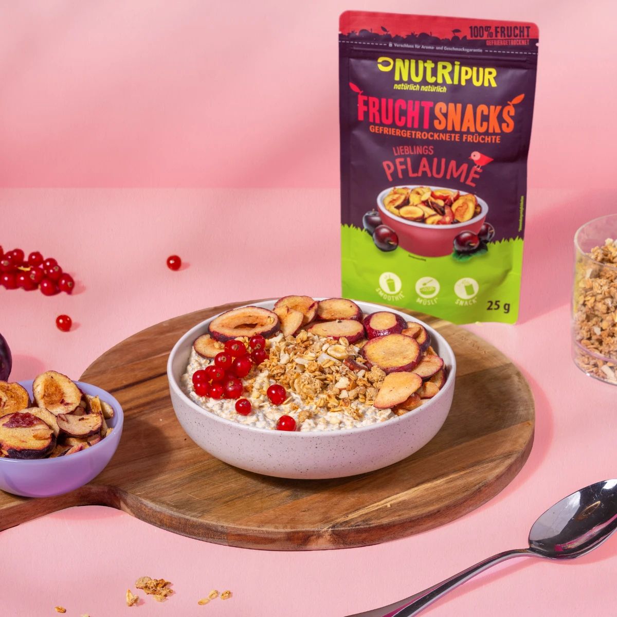 Beutel mit gefriergetrockneten Pflaumen-Snacks. Auf Holzbrett mit Schüssel Müsli, Beeren und Pflaumen. Glas mit Müsli.