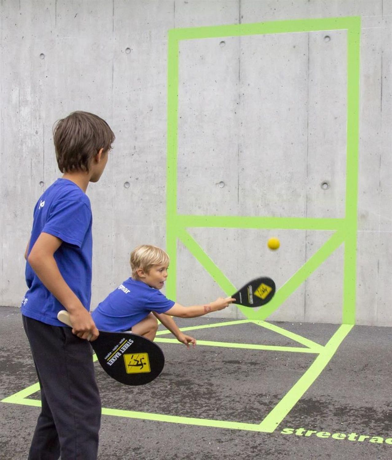 Zwei Kinder spielen Street Racket. Einer schlägt den Ball mit einem schwarzen Schläger. Auf dem Boden ist ein grünes Feld markiert.
