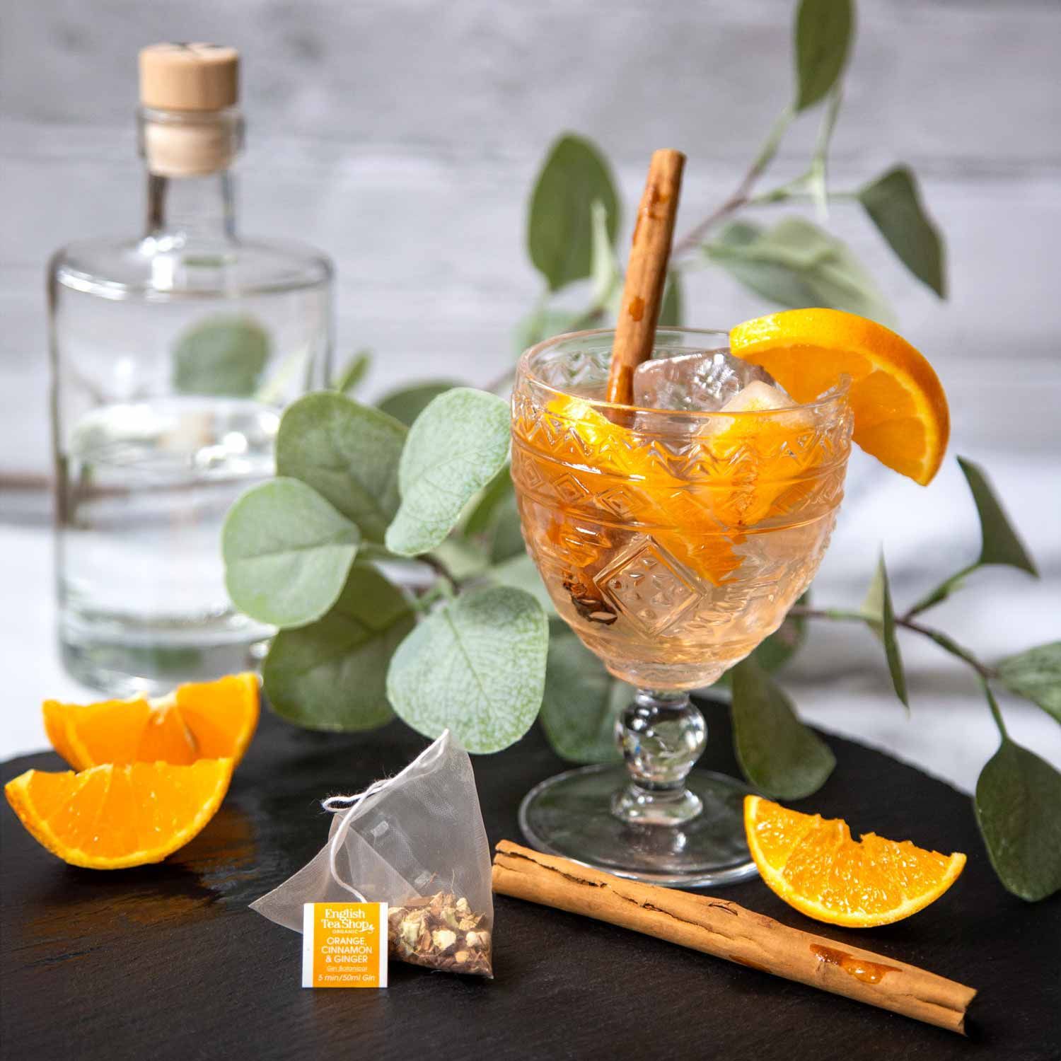 Glas mit Gin-Cocktail. Orange, Zimtstange, Eiswürfel. Teebeutel. Gin-Flasche im Hintergrund.