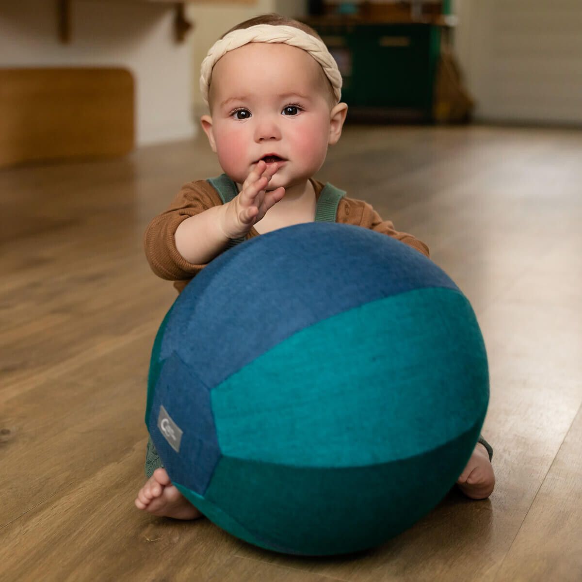 Baby sitzt mit einer blauen und grünen Stoffkugel. Das Kind hat einen Stirnband und hebt die Hand.
