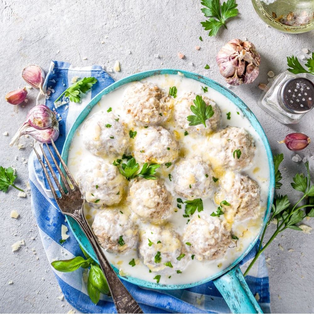 Pfanne mit Köttbullar in Sauce, Knoblauchzehen, Kräutern und Salzstreuer. Blaue Serviette.