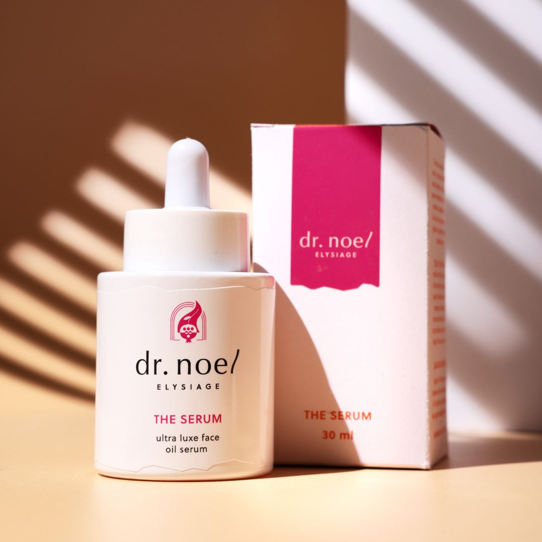 Weiße Serumflasche und Verpackung. Aufschrift: dr. noel/ ELYSIAGE, THE SERUM, ultra luxe face oil serum. Rosa Akzente. Hintergrund: hellbraun.